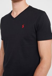 Muž oblečený v černém tričku s výstřihem do V a krátkým rukávem, se malým červeným vyšitým logem polo hráče na levé části hrudi.