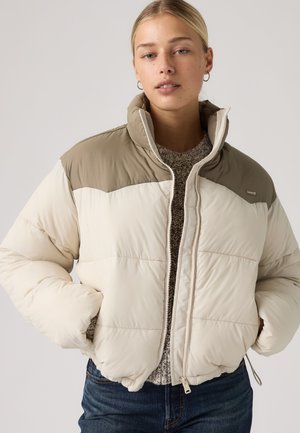 Jeune femme portant une doudoune beige et marron clair, les mains dans les poches, debout devant un fond gris uni.