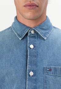 Lichtblauwe denimshirt met een klassieke kraag, voorzien van zilveren knopen en een borstzak met een klein rood, wit en blauw logo.