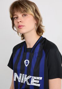 Nike jersey in black, featuring vertical blue stripes. Includes a collar, large white logo, and round emblem centered above the logo.