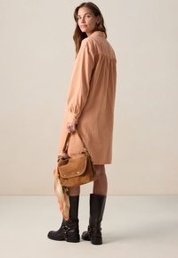 Robe chemise de couleur pêche avec une coupe ample, des manches longues et des poignets boutonnés. Sac à main en daim marron avec une écharpe à motifs et des bottes noires à midi.