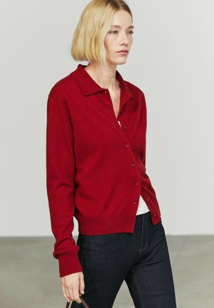 Femme aux cheveux blonds courts portant un cardigan rouge boutonné et un jean foncé sur un fond clair uni.