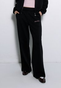 Schwarze Sweatpants mit weitem Bein-Design, aus einem weichen Stoff gefertigt, mit einem bestickten rosa Logo und seitlichen Taschen.