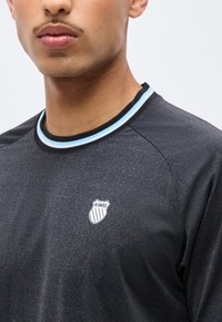 Κοντινό πλάνο ενός ατόμου που φοράει μπλουζάκι K-Swiss σκούρου γκρι με γιακά ριγέ σε μαύρο, λευκό και μπλε.