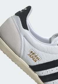 Bílá kožená teniska s černými semišovými doplňky, třemi černými pruhy a zlatým logem "adidas JAPAN". Texturovaná podrážka a prošité detaily.