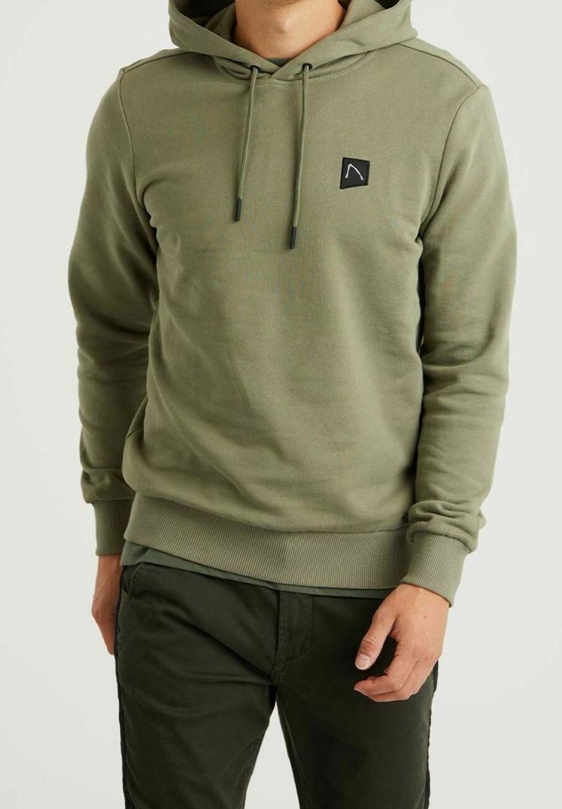 Homme portant un sweat à capuche vert olive avec cordons et un pantalon noir, debout devant un fond clair uni.