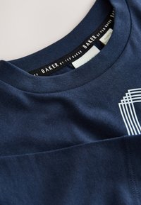 Laivastonsininen pyöreäkauluksinen paita, jossa musta sisäkaulusetiketti, jossa lukee "BAKER by Ted Baker", ja valkoinen geomeetrinen painatus, osittain näkyvissä rinnassa.