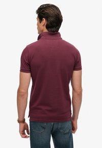 Bordeauxrode polo met een geribbelde textuur, korte mouwen en een opgevouwen kraag, gecombineerd met een blauwe spijkerbroek, gezien vanaf de achterkant.