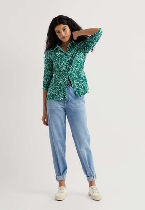 Grünes, langärmliges Hemd mit Blumenmuster, kombiniert mit hellblauen Jeans mit aufgerolltem Saum und weißen Sneakers, getragen von einem Model vor neutralem Hintergrund.