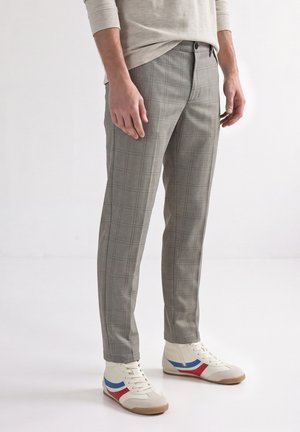 Mann trägt slim-fit graue karierte Hose und weiße High-Top-Sneaker mit roten und blauen Streifen auf weißem Hintergrund.
