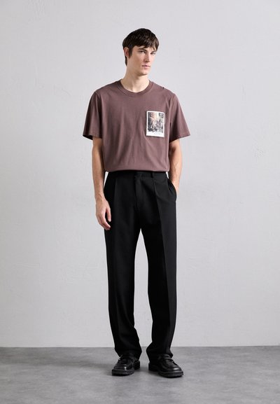 Barna pamut póló, mellzsebes nyomattal, fekete, bő szabású nadrággal és fekete bőr cipővel párosítva. Egyszínű háttér.