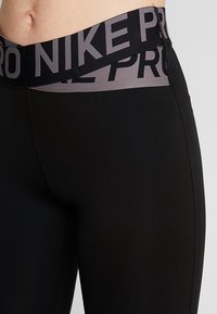 Leggings de sport noirs avec une large ceinture élastique grise sur laquelle est imprimé "NIKE PRO". Texture lisse et design ajusté.
