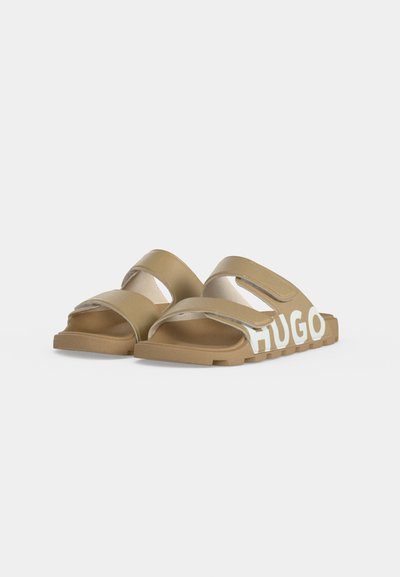 Paire de sandales slides marron clair avec deux brides ajustables et marquage blanc "HUGO" sur le côté de la semelle.
