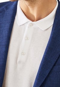 Biała koszulka polo z trzema guzikami, noszona pod niebieską, teksturowaną marynarką. Kołnierzyk jest płaski, a tkaniny wydają się lekkie.