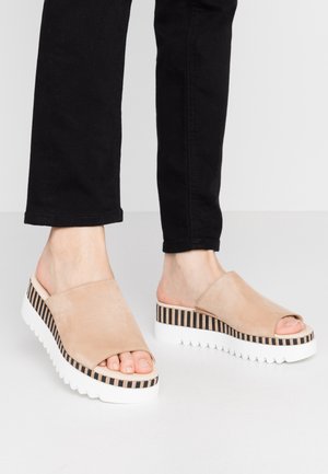 Sandales à glisser en suède beige avec un bout ouvert, dotées d'une plateforme rayée à motifs et d'une épaisse semelle blanche. Pantalons noirs avec un ourlet légèrement raccourci.