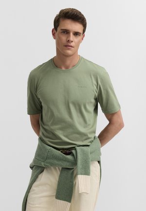 Junger Mann mit kurzen braunen Haaren, der ein hellgrünes T-Shirt und einen grünen Strickpullover trägt, der um seine Taille gebunden ist, vor einem einfarbigen Hintergrund.