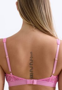 Rosa Spitzen-BH mit verstellbaren Trägern und Haken- und Ösenverschluss. Auffälliges Tattoo mit Ziffern auf dem Rücken sichtbar.