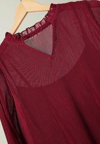 Blouse plissée bordeaux avec un décolleté en V et un col à volants, en tissu transparent, présentant des rayures verticales et une silhouette fluide.