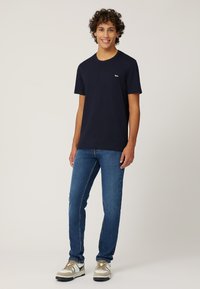 Marineblauw katoenen t-shirt met korte mouwen, voorzien van een klein logo op de borst. Gecombineerd met donkerblauwe jeans en sneakers met beige accenten.