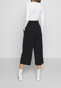 Pantalons noirs à jambes larges avec une taille haute, dotés de poches latérales. Portés avec un haut blanc à manches longues et des bottes blanches arrivant aux chevilles.