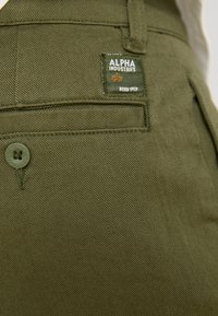 Olive rohelised püksid siledast materjalist, väikese Alpha Industries sildiga, nööp kinnitusega ja diagonaalses tasku disainiga.