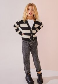 Schwarz-creamfarbener Streifenstrickjacke mit V-Ausschnitt und Knopfverschluss, kombiniert mit hochgeschnittenen grauen Jeans und schwarzen Ankle Boots.