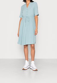 Kvinna i en ljusblå knälång klänning med knappar, uppvikt ärm, midjeband och vita högklackade platåskor.