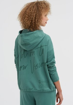 Frau mit lockigem Haar, die einen türkisfarbenen Kapuzenpullover und eine Hose trägt, seitlich gedreht und den Text auf der Rückseite des Pullovers zeigend.