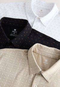Tres camisas de botón de ajuste medio, en blanco, negro y beige, dobladas y con pequeños diseños geométricos y etiquetas de marca visibles.
