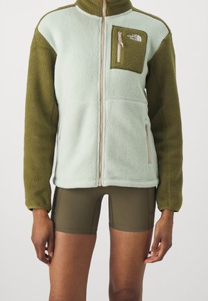 Person, die eine zweifarbige grüne Fleecejacke mit Reißverschlusstasche an der Brust und olivgrüne Fahrradshorts vor schlichter Hintergrund trägt.