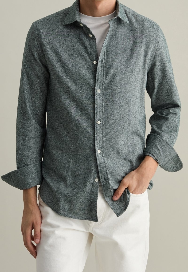 Camicia button-up di colore verde scuro, in tessuto testurizzato. Presenta un colletto, bottoni e maniche arrotolate. Abbinata a pantaloni bianchi per un contrasto.