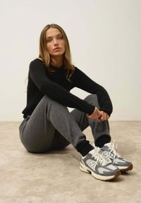 Svart ribbad tröja, grå sweatpants och grå sportskor med vita accenter. Sittande pose på en jämn yta med en neutral bakgrund.