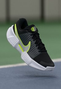 Μαύρο αθλητικό παπούτσι με νέον πράσινες λεπτομέρειες, υφασμάτινη επιφάνεια, λευκή σόλα και ισοροπημένος σχεδιασμός για σταθερότητα και υποστήριξη.