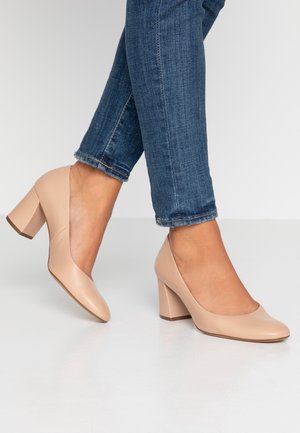 Béžové kožené lodičky s kulatou špičkou a masivním blokovým podpatkem. Nosené s tmavě modrými džíny, které ukazují hladkou, lesklou texturu.