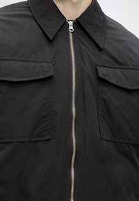 Μαύρο μπουφάν με φερμουάρ, με μυτερό γιακά και δύο μεγάλες τσέπες με καπάκι στο στήθος, που φοριέται από άτομο με ορατό λαιμό και γραμμή σαγονιού.