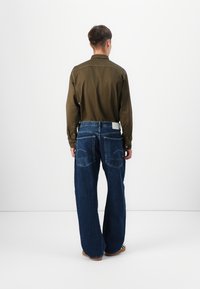 Chemise à manches longues vert olive avec des détails de boutons, associée à un jean en denim bleu ample avec de grandes poches arrière et une étiquette blanche.