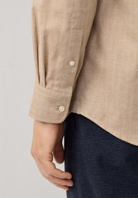 Close-up van een beige linnen shirt met een knoopsluiting bij de manchetten, textuurstof en een getailleerde pasvorm, met subtiele plooien nabij de mouwen.