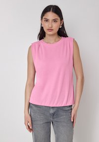 Top senza maniche rosa in tessuto morbido con scollatura rotonda e pieghe sottili sulle spalle, abbinato a jeans in denim grigio.