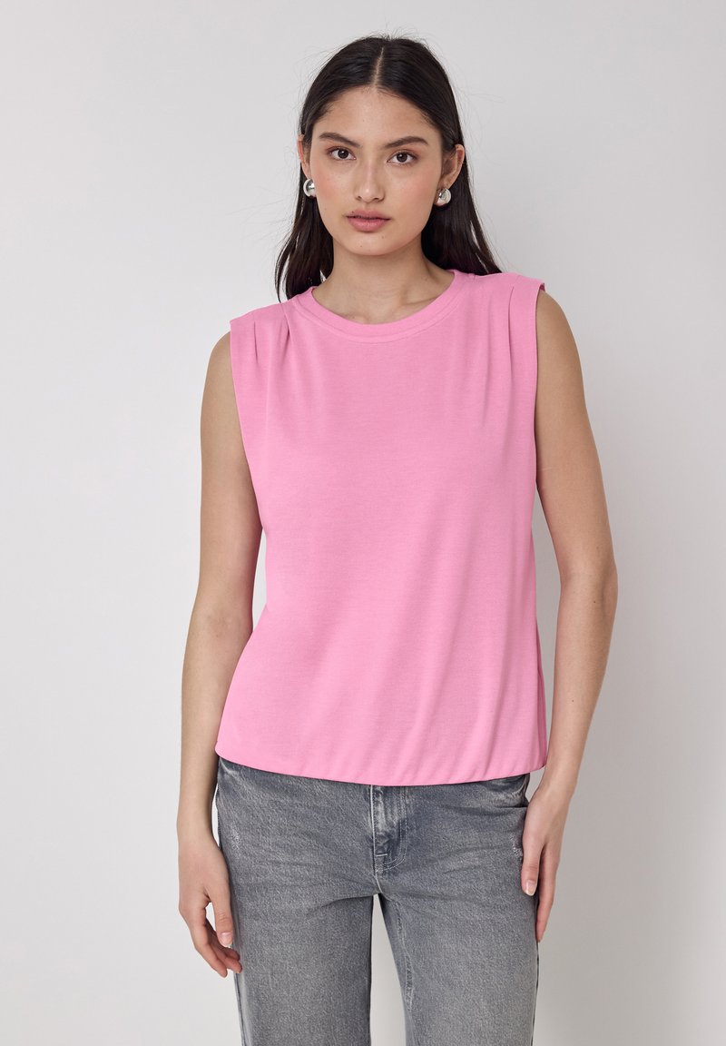 Top senza maniche rosa in tessuto morbido con scollatura rotonda e pieghe sottili sulle spalle, abbinato a jeans in denim grigio.
