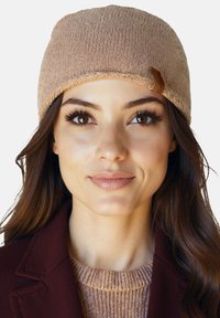 Hosszú barna hajú nő, aki bézs kötött sapkát, bézs pulóvert és bordó kabátot visel, előre néz semleges arckifejezéssel.