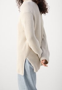 Gebreide trui in beige, met een relaxte pasvorm, geribbelde textuur, zijsplitten en lange mouwen. Gecombineerd met lichtblauwe jeans.