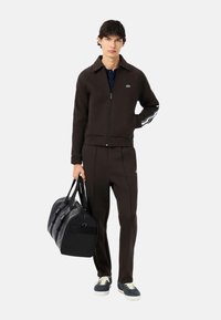 Giacca sportiva marrone con cerniera e logo verde, pantaloni abbinati e borsa da viaggio nera. Scarpe da ginnastica con suole bianche, lacci chiari e dettagli blu.