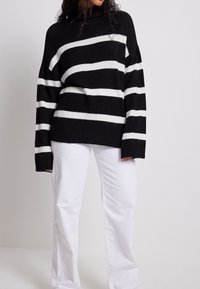 Gestreifter schwarz-weißer Strickpullover mit hohem Kragen, voluminösen Ärmeln und gerippten Bündchen, kombiniert mit weißen, weit geschnittenen Hosen.