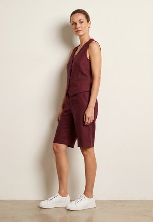 Žena stojící opřená o zeď, oblečená v bezrukávovém bordovém vestě na knoflíky, odpovídajících kraťasech po kolena a bílých teniskách.