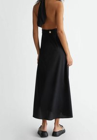 Schwarzes, rückenfreies Maxikleid mit Halterneck und Sternakzent in der Taille. Glatte Textur und bodenlanger Schnitt.