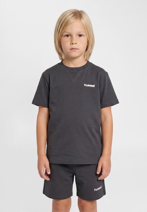 Blond dziecko stoi, nosi ciemnoszary krótki rękaw i pasujące szorty z logo "hummel" na piersi i nodze.