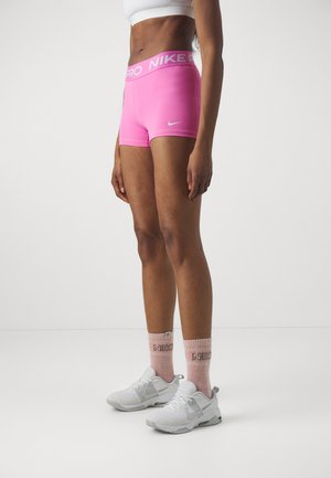 Shorts Nike Pro roses avec une large taille, associés à des baskets blanches et des chaussettes roses claires avec le texte 'Nike'. Materiau lisse et extensible.