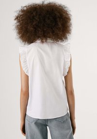 White sleeveless blouse with ruffled shoulder accents, soft cotton fabric, and a relaxed fit. Back view shows a rounded hem and subtle texture.