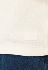 Λευκό υφασμάτινο σχέδιο με υφή και μοτίβο πλέγματος, με μια μικρή ανάγλυφη ετικέτα Vans στο τελείωμα, σε συνδυασμό με σκούρο τζιν από κάτω.