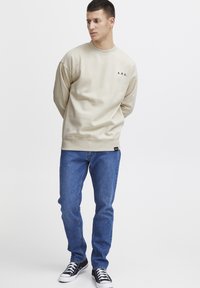 Sudadera beige con cuello redondo y mangas acampanadas, que presenta un pequeño logo negro "A.R.C.". Combinada con vaqueros azules y zapatillas negras.
